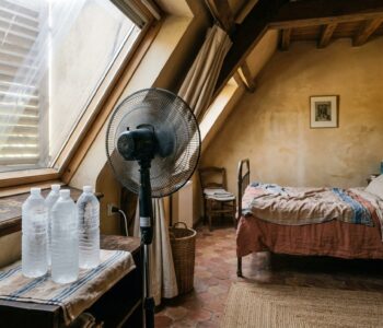 Un ventilateur sur pied dirigé vers des bouteilles d'eau gelées posées devant une fenêtre calfeutrée avec un panneau de